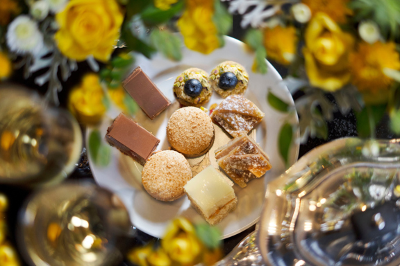 Plate of assorted high-tea pastries at Prestonfield House, Edinburgh, featuring chocolate slices, coconut macaroons, pistachio and blueberry bites, shortbread and layered sweet treats, surrounded by fresh yellow flowers and sparkling glassware — a refined high tea in Edinburgh